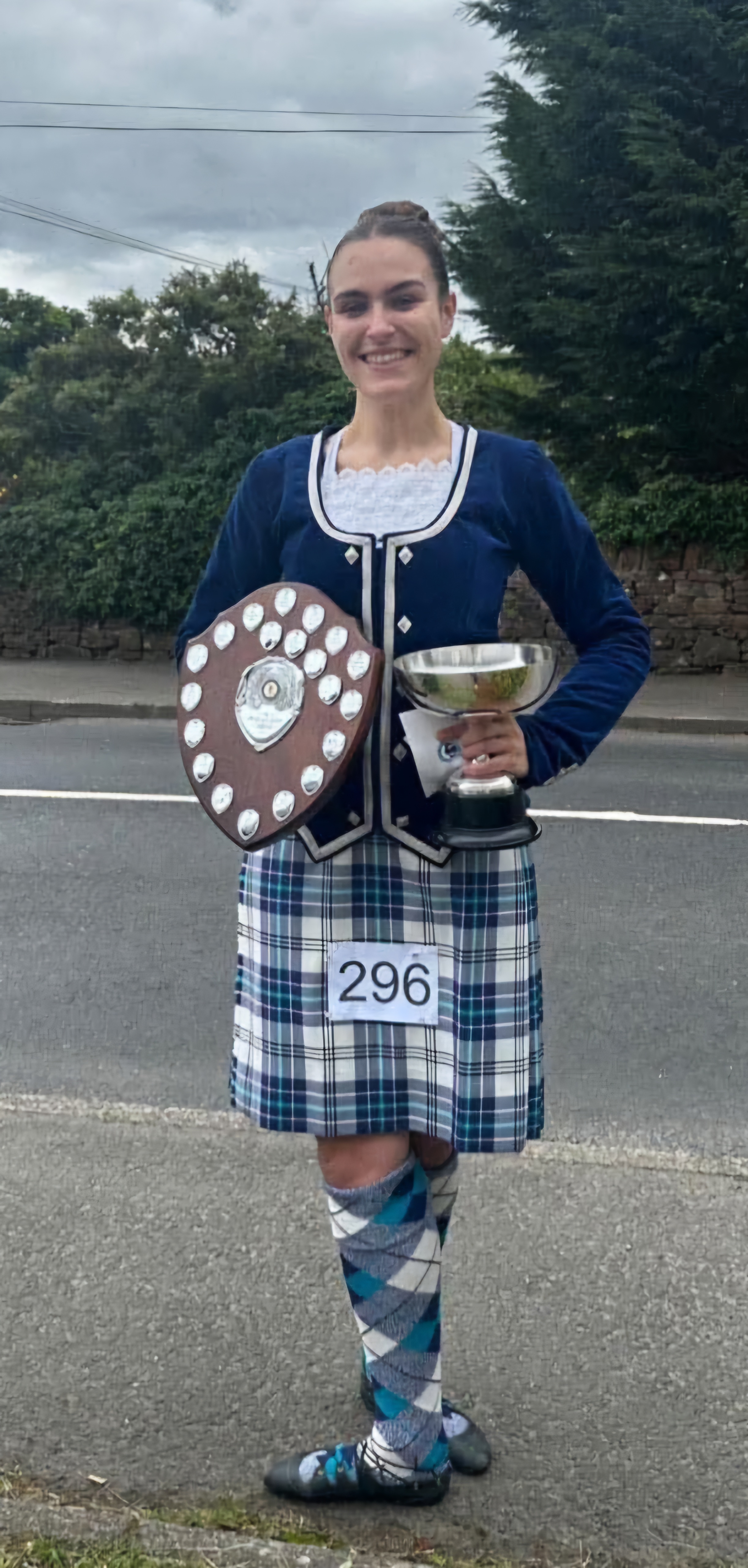 Miss Nadia dressed in tartan, holding a silver cup and a silver shield covered in awards