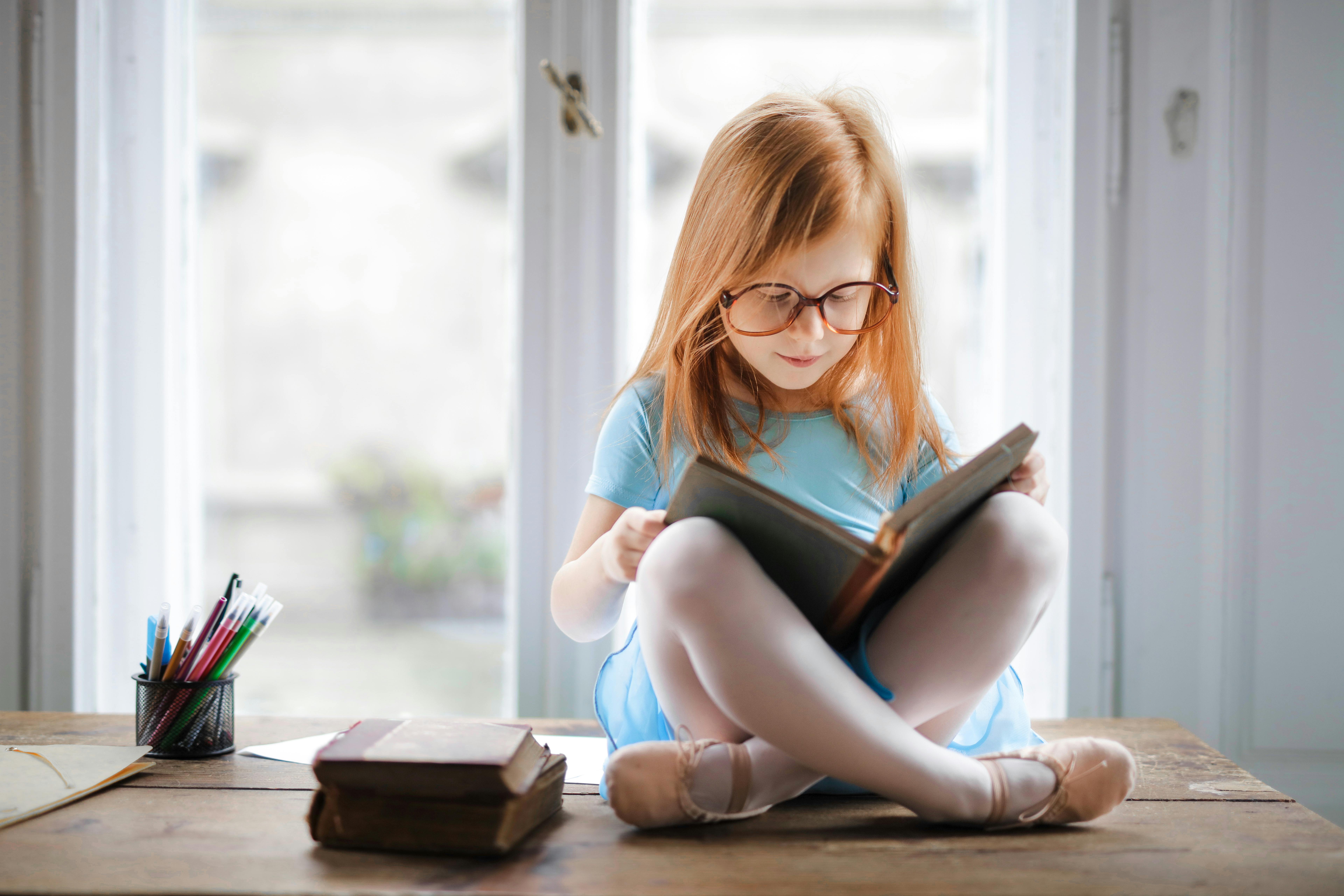A yong girl wearing ballet slippers sits cross-legged, reading a weighty tome